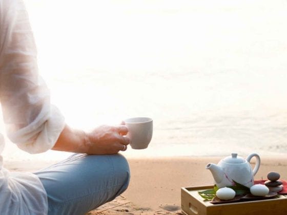 Eine Frau hält am Strand eine Tasse mit Tee aus dem Ayurveda.