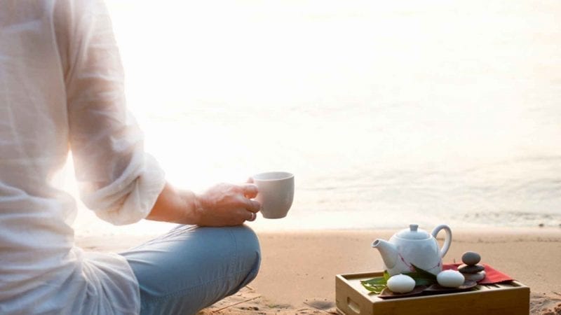 Eine Frau hält am Strand eine Tasse mit Tee aus dem Ayurveda.